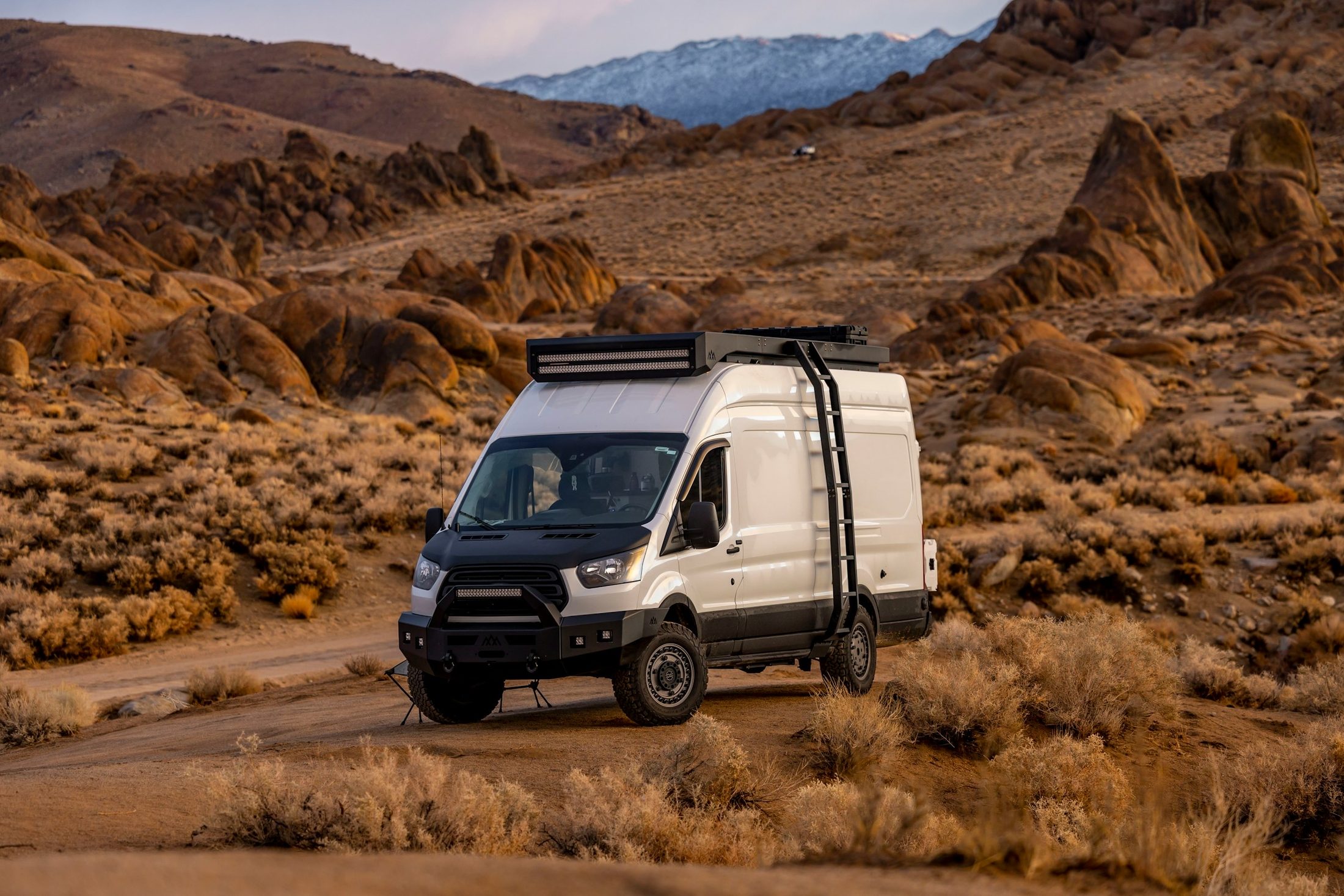 A white Ford Transit camper van with a roof rack parked among the Alabama Hills boulders at golden hour